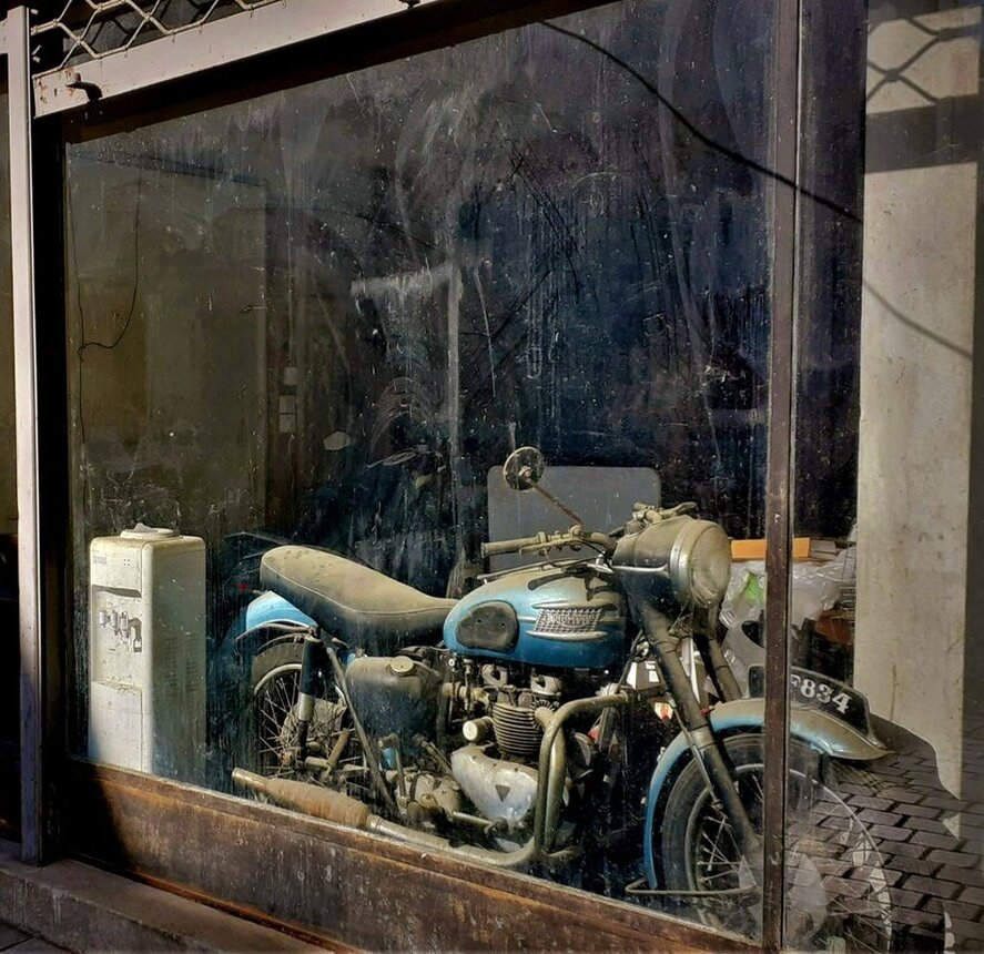 An elderly Triumph motorbike looking new aside from years of dust, sitting in a deserted looking shop front. Nicosia, CY